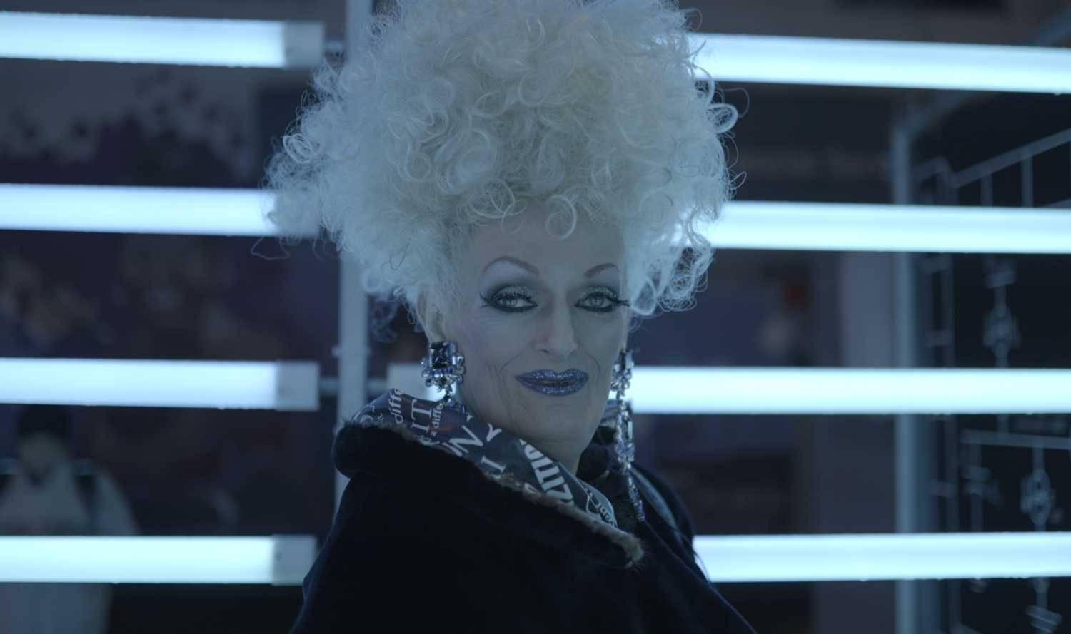 Empress of Vancouver B %2B W Glamourous woman with glittery makeup, an updo and large dangling earrings wearing a fur lined, collared coat. Drag Icon Oliv Howe.